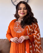 Woman wearing an orange traditional outfit with intricate patterns, standing indoors.