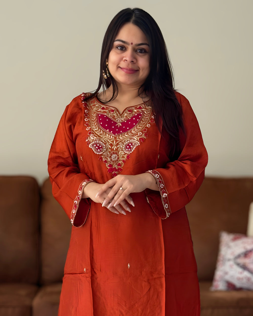 Woman wearing an orange embroidered traditional outfit standing in a room with a brown sofa.