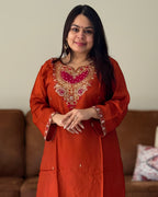 Woman wearing an orange embroidered traditional outfit standing in a room with a brown sofa.