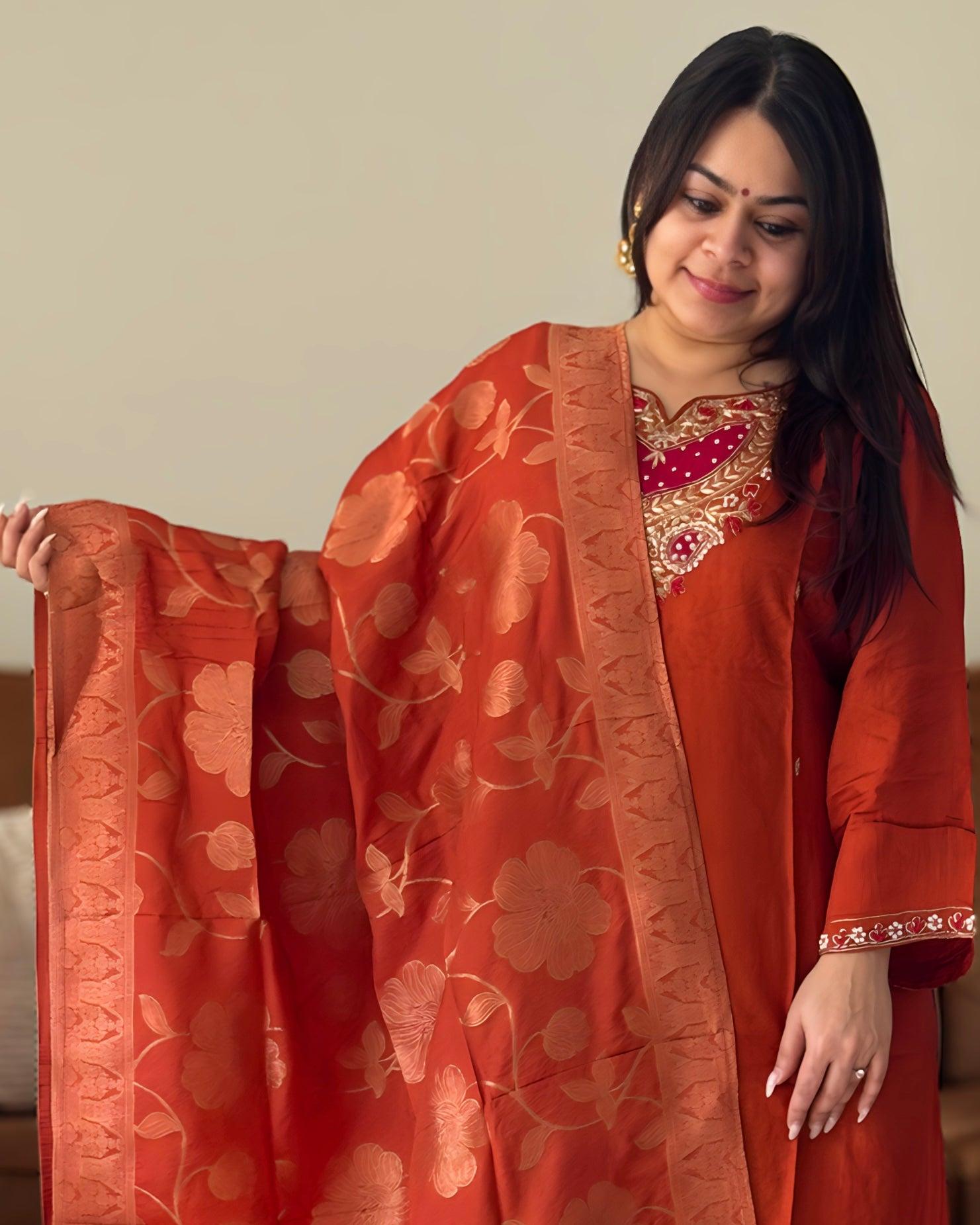 Woman wearing a traditional outfit with an orange dupatta, standing against a plain background.