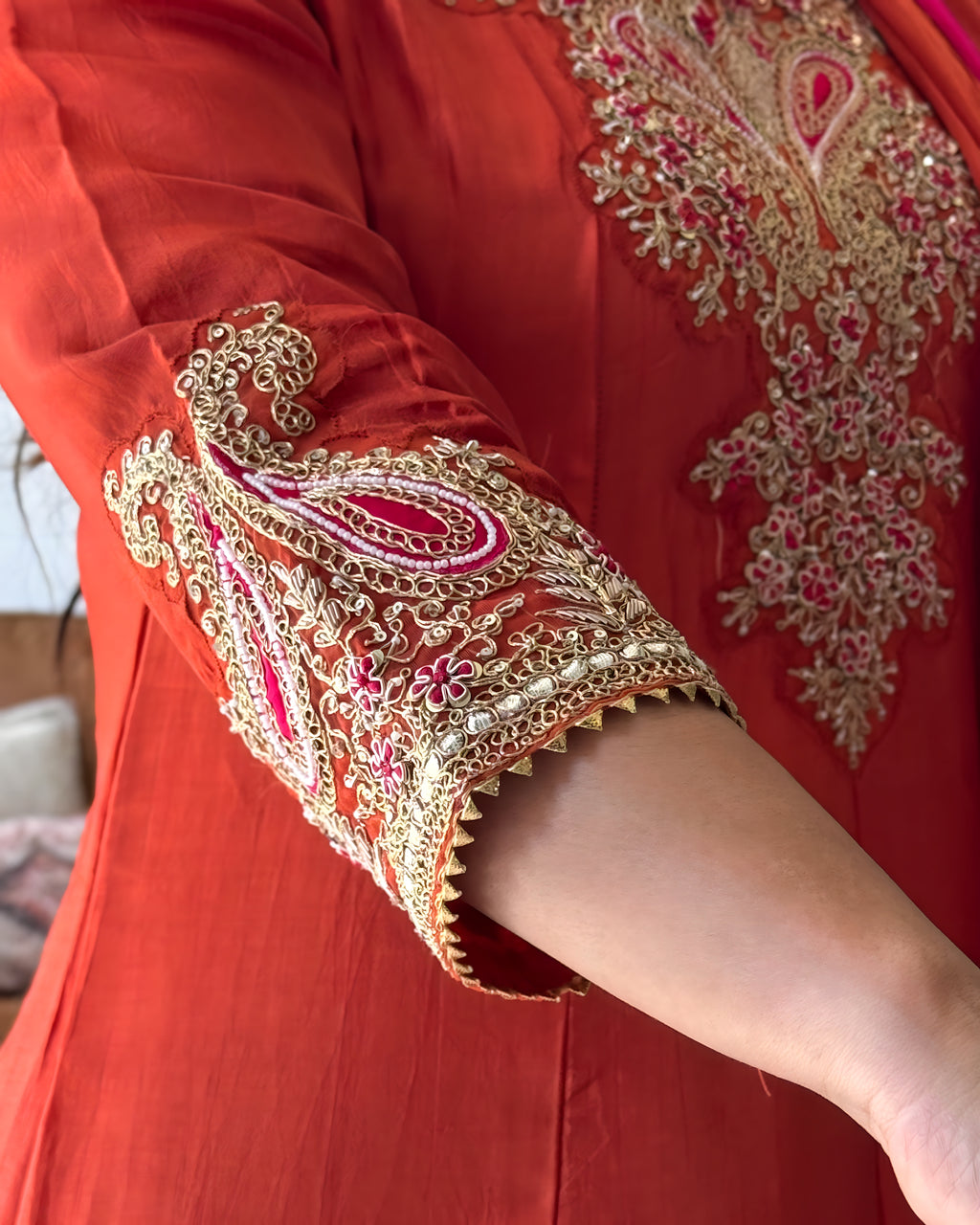 Close-up of a red traditional outfit with ornate gold embroidery and pink accents on the sleeve and chest area, worn by a person whose arm is visible.