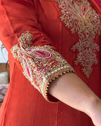 Close-up of a red traditional outfit with ornate gold embroidery and pink accents on the sleeve and chest area, worn by a person whose arm is visible.