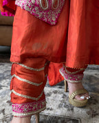 Close-up of a person wearing a bright orange-red traditional outfit with pink and gold embroidered details, paired with gold platform sandals, standing on a textured rug.