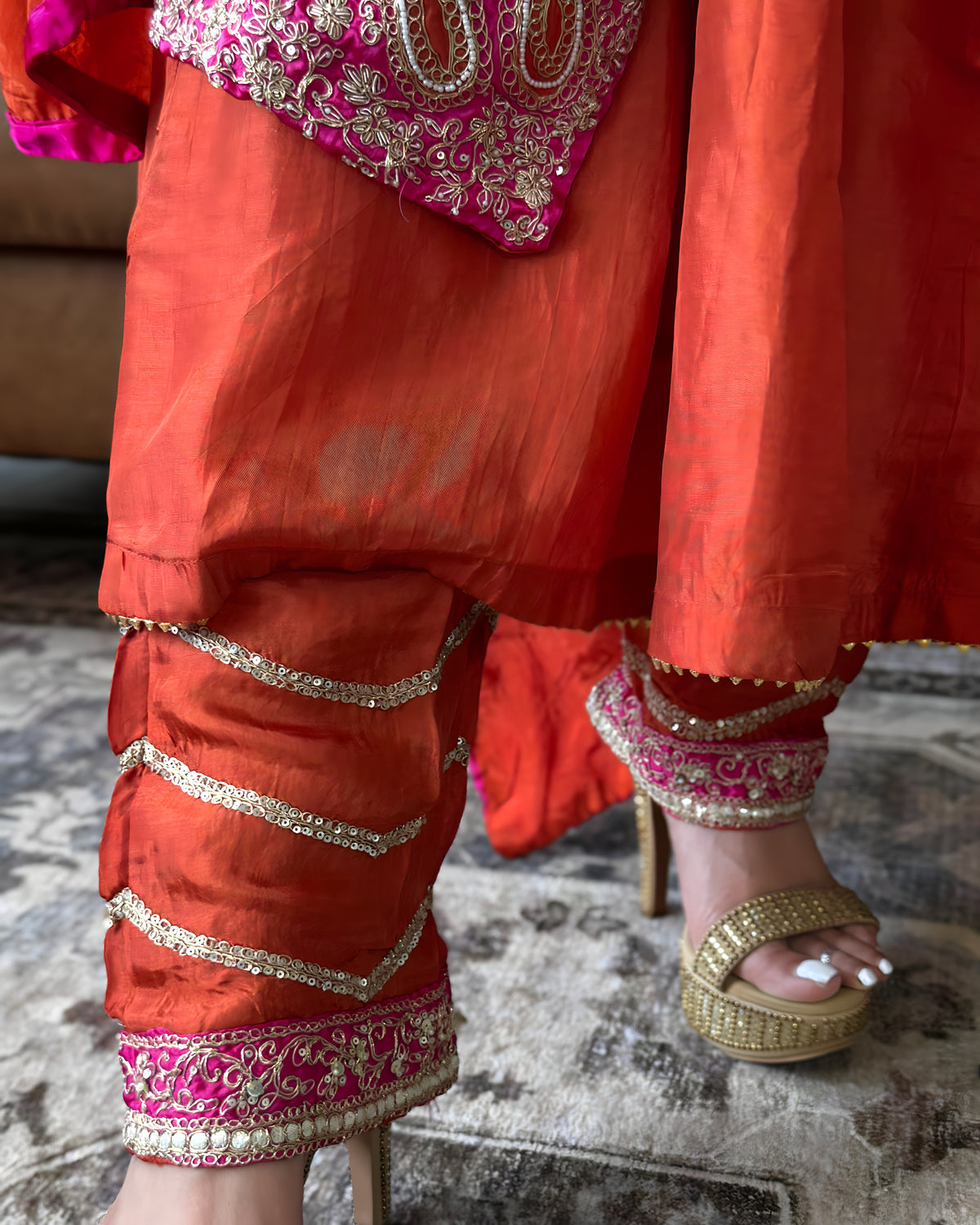 Close-up of a person wearing a bright orange-red traditional outfit with pink and gold embroidered details, paired with gold platform sandals, standing on a textured rug.