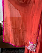 A close-up of a coral orange-red sheer fabric, likely a dupatta or sari, featuring a vibrant magenta border and white floral embroidery with metallic embellishments at the bottom corner.