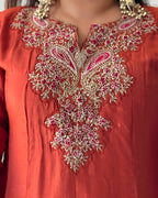 Close-up of a coral orange traditional garment with elaborate gold embroidery and pink floral details around the neckline, worn with gold jewelry.