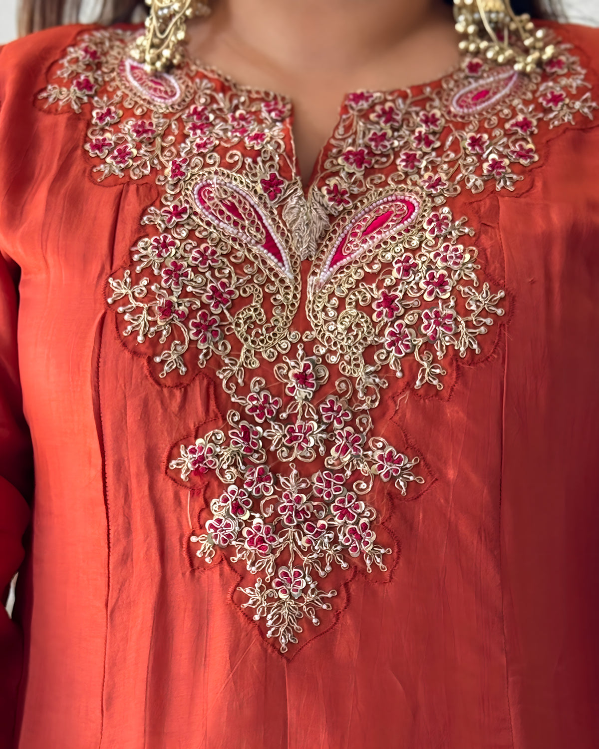 Close-up of a coral orange traditional garment with elaborate gold embroidery and pink floral details around the neckline, worn with gold jewelry.