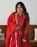A woman wearing a red traditional outfit with gold and pink border details, standing in front of a brown sofa against a plain light wall.