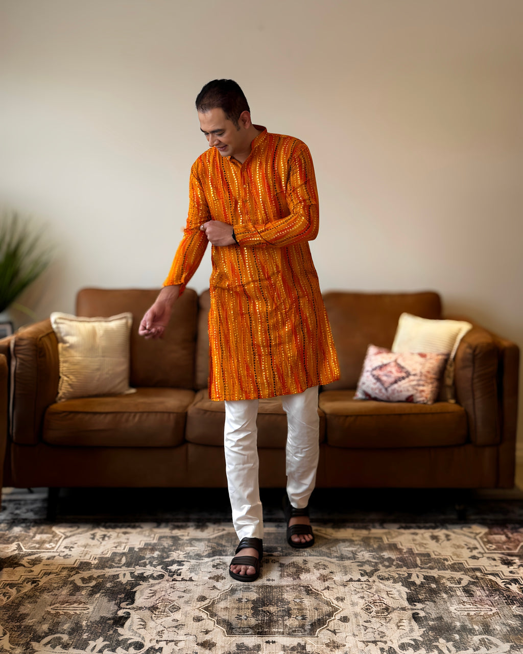 Man wearing an orange kurta with white pants standing in a living room.
