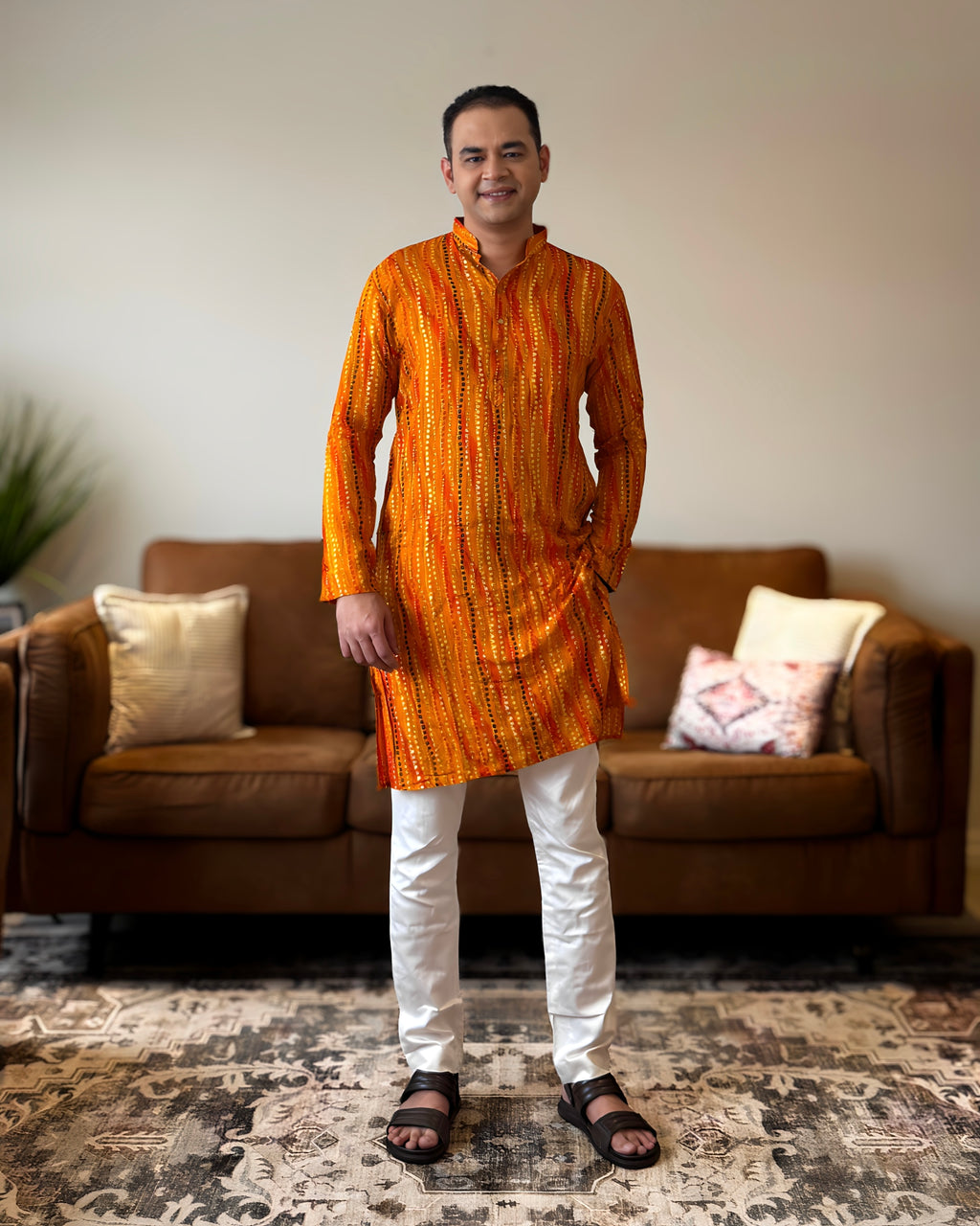 Man wearing an orange kurta with white pants standing in a living room.