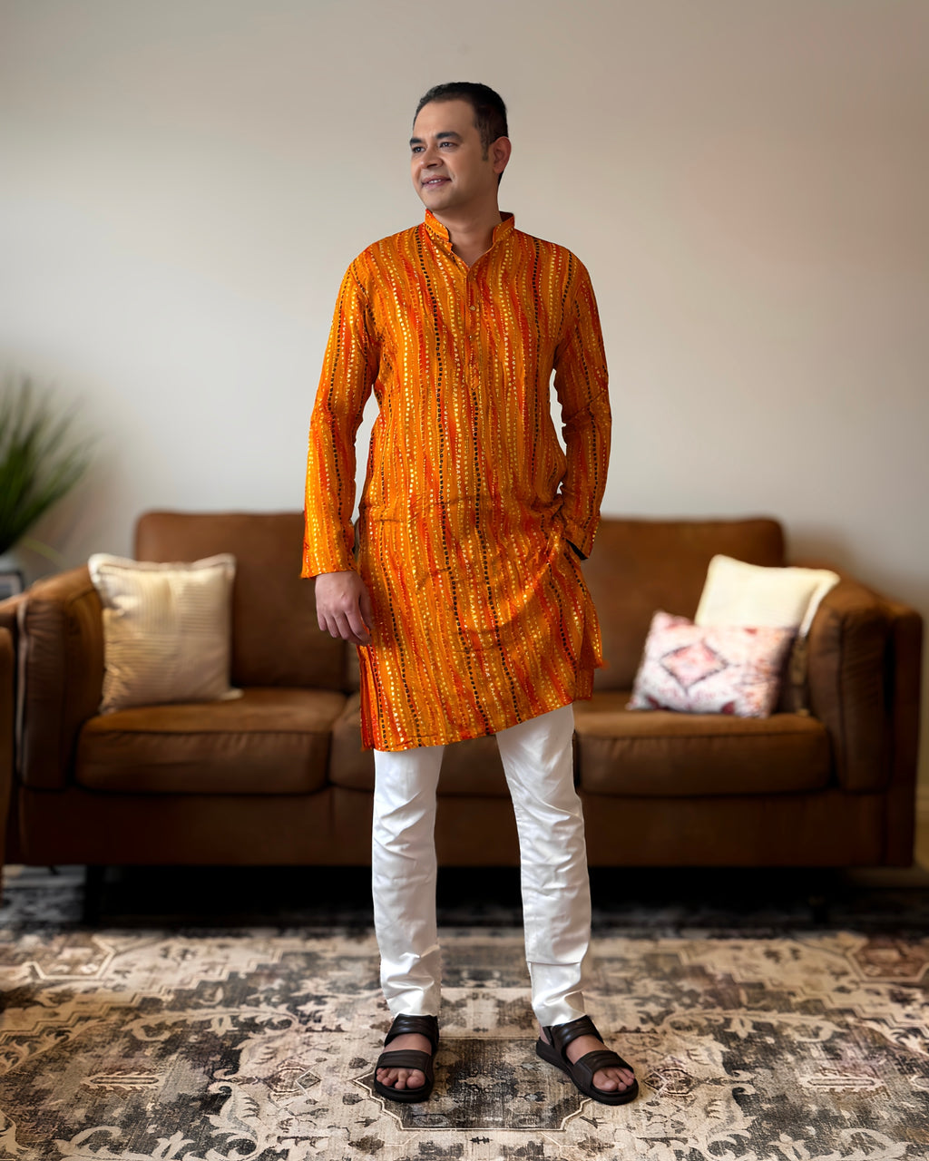 Man wearing an orange kurta with white pants standing in a living room.