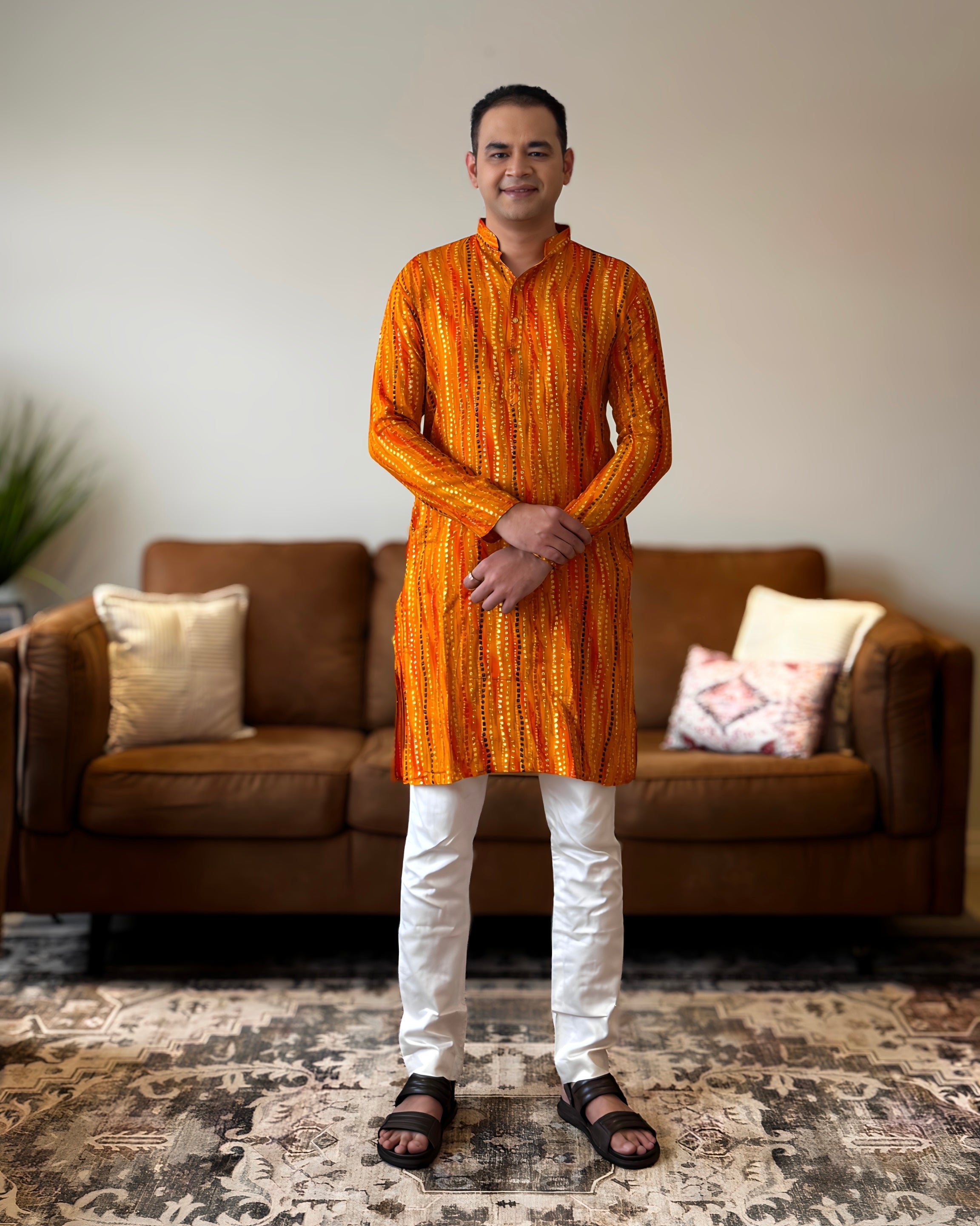 Man wearing an orange kurta with white pants standing in a living room.