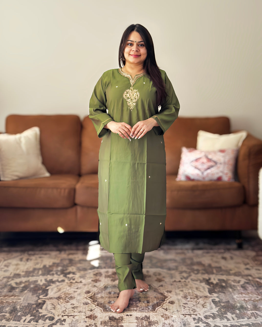 Woman in a green traditional outfit standing in a living room with a brown sofa and patterned rug.