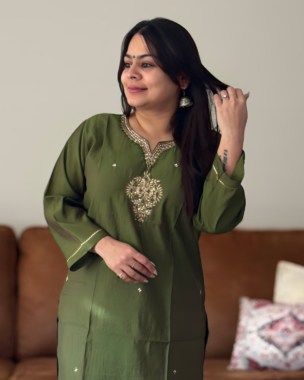 Woman in a green traditional outfit with embroidery, standing in a room with a brown couch and patterned pillow.