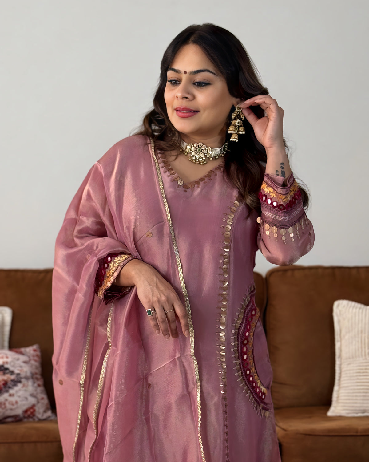 A woman wearing a dusty pink embroidered kurta with gold metallic borders and embellishments, styled with a matching dupatta and traditional jewelry, standing in front of a brown sofa.