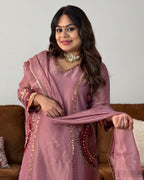A woman wearing a dusty pink traditional outfit with gold embroidery and a matching dupatta, standing in front of a neutral wall and a brown sofa.