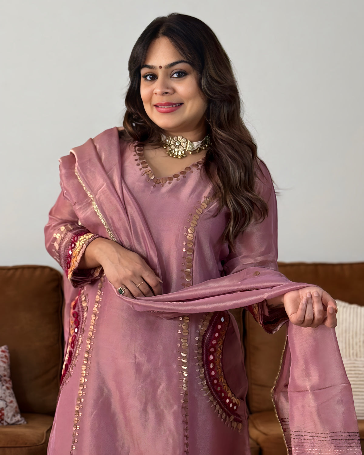 A woman wearing a dusty pink traditional outfit with gold embroidery and a matching dupatta, standing in front of a neutral wall and a brown sofa.