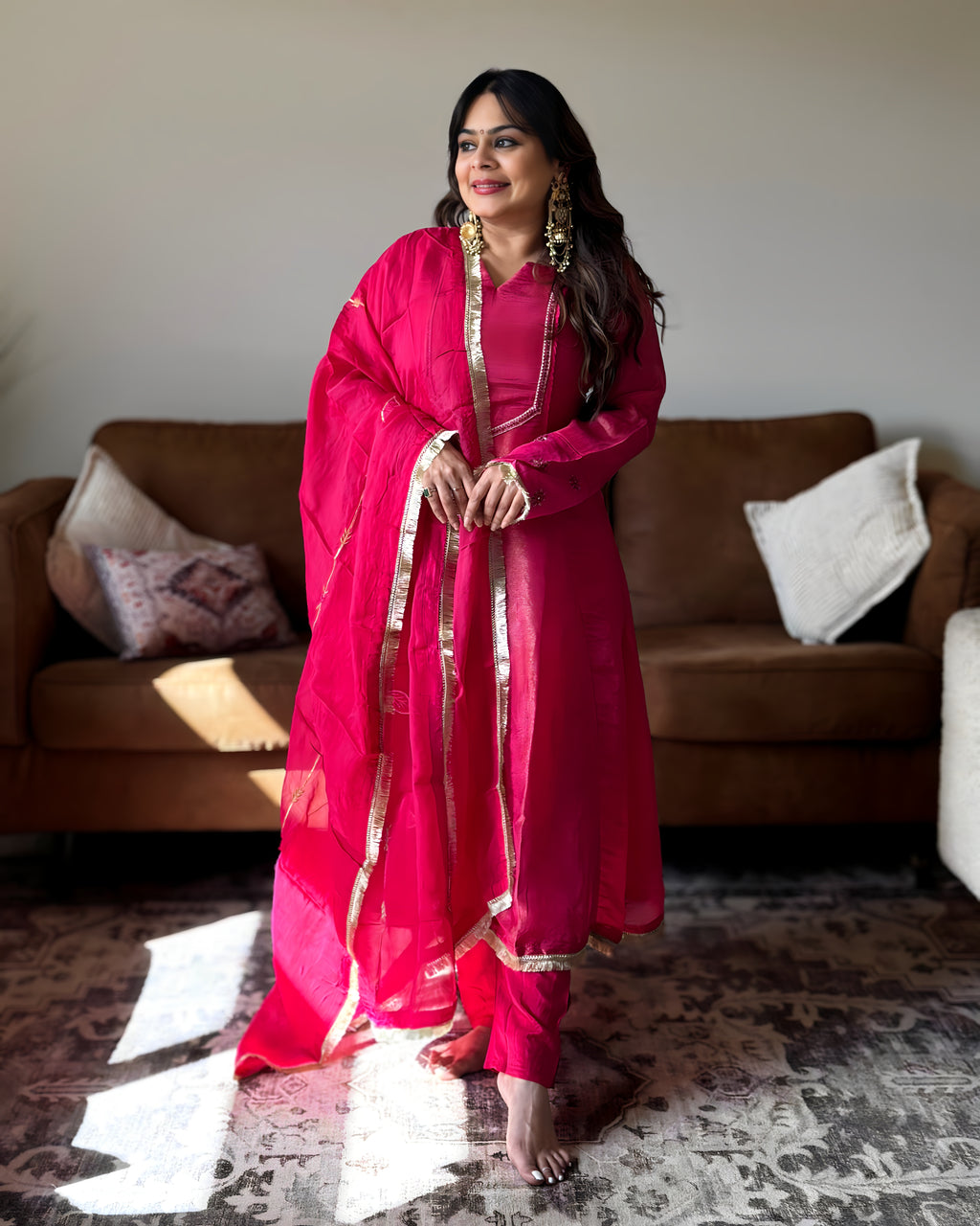 A woman wearing a bright pink traditional outfit with gold trim stands barefoot in a living room in front of a brown sofa.