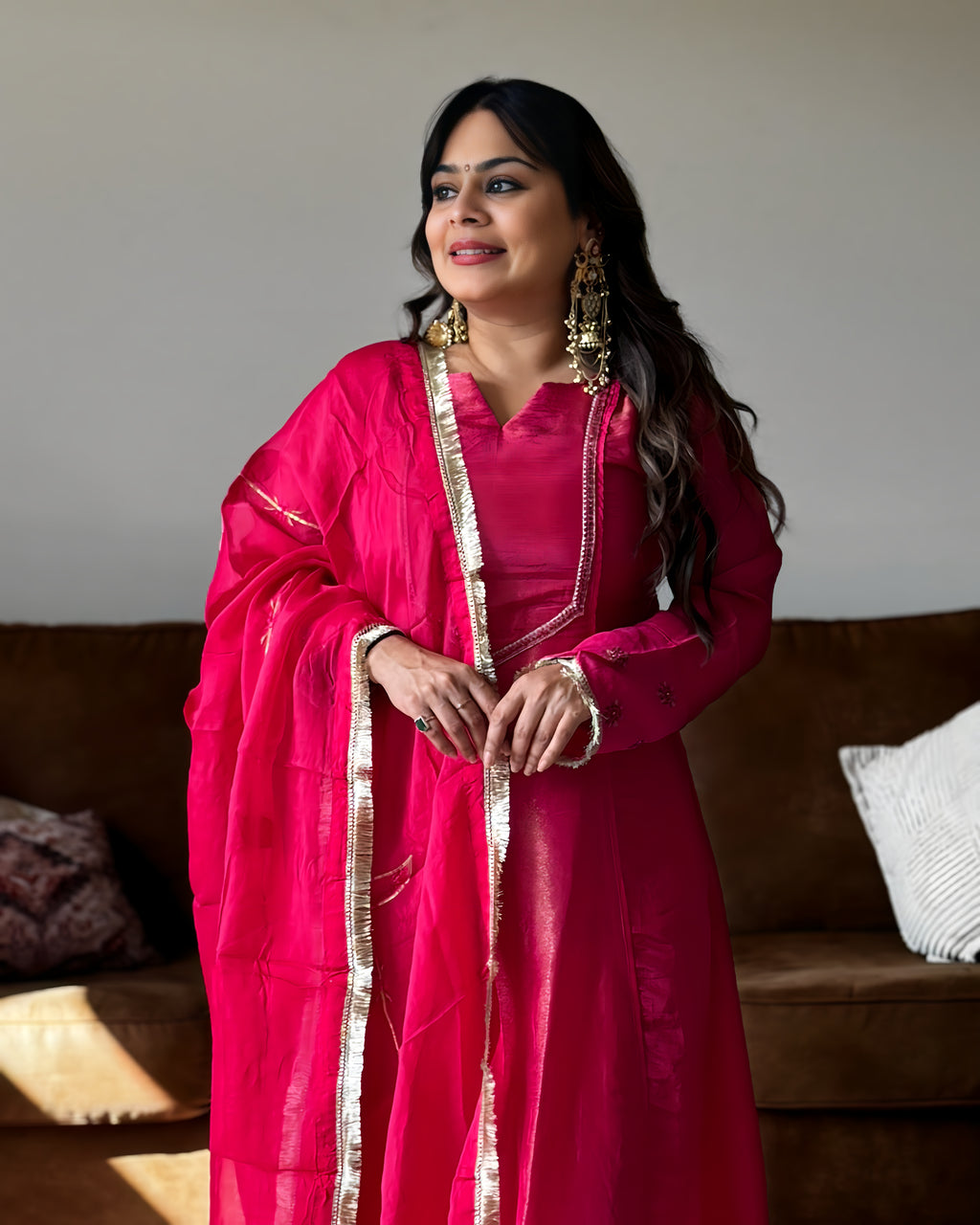 A woman wearing a bright pink traditional outfit with gold trim and a matching sheer dupatta, standing indoors in front of a brown sofa.