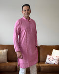 Man wearing a pink kurta standing in a living room with a brown sofa and decorative pillows.