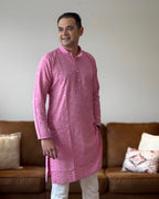 Man wearing a pink kurta standing in a room with a brown couch and decorative pillows.