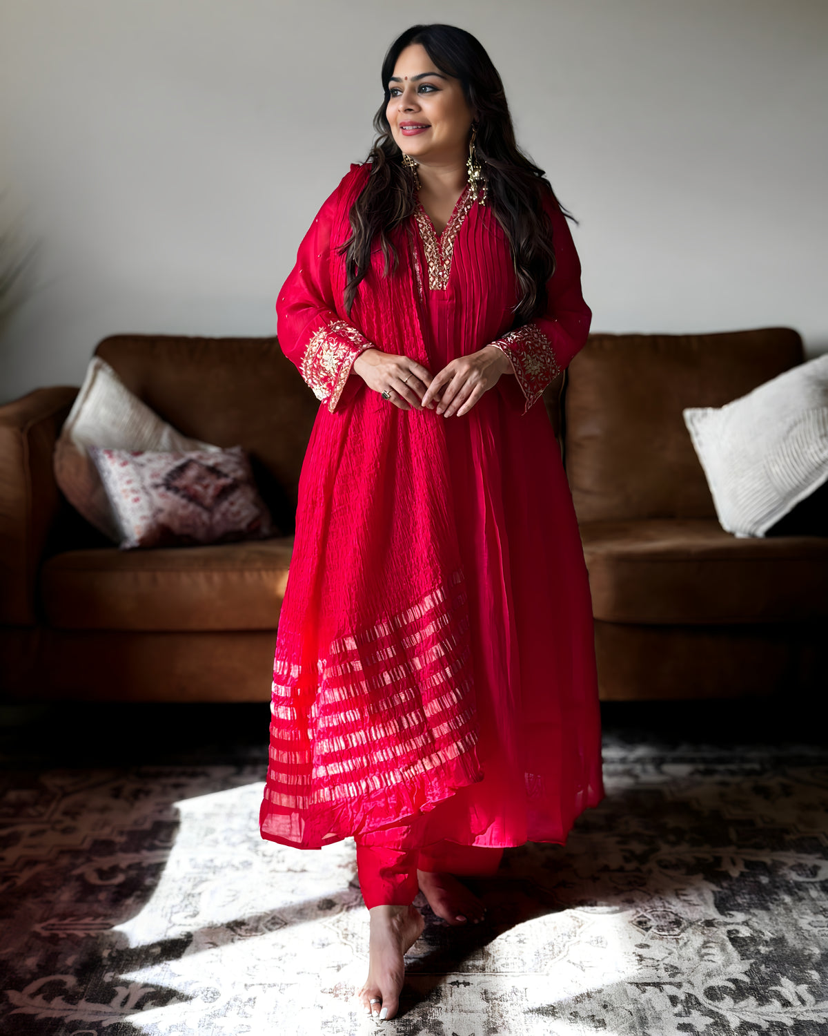 A woman stands barefoot in a living room wearing a bright red traditional outfit with gold embroidery on the neckline, sleeves, and hem. A brown sofa and patterned rug are visible in the background.