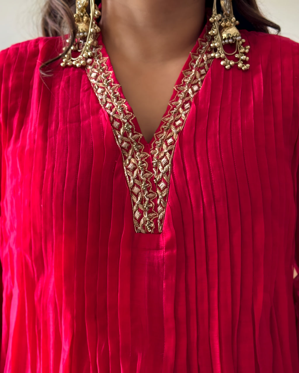 A close-up of a bright pink pleated fabric garment with ornate gold embroidery around a split V-neckline, worn by a model with large gold statement earrings.