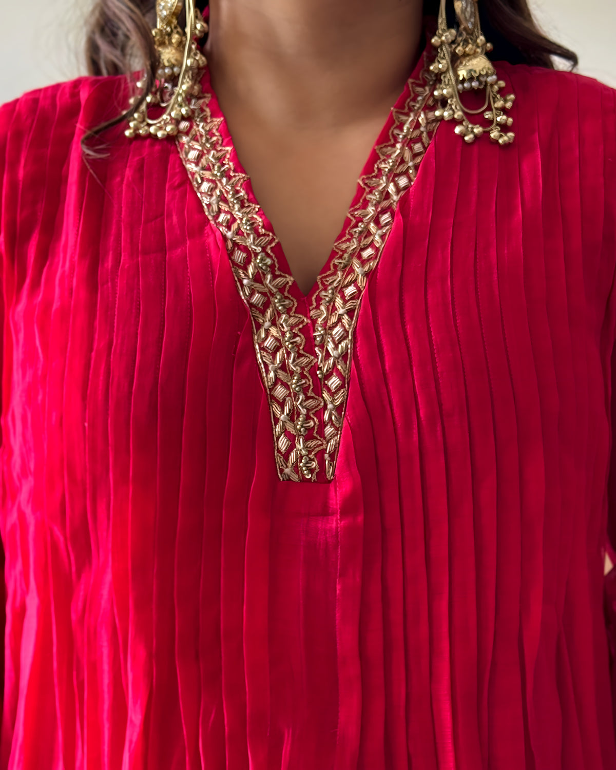 A close-up of a bright pink pleated fabric garment with ornate gold embroidery around a split V-neckline, worn by a model with large gold statement earrings.