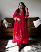 A woman wearing a vibrant red traditional Indian outfit with gold embroidery and a matching dupatta, standing in a living room in front of a brown sofa.