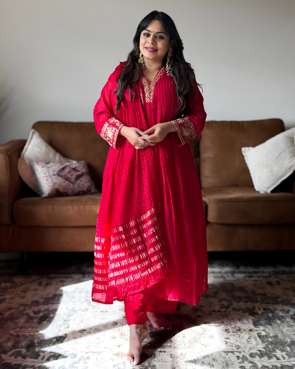 A woman modeling a bright red traditional Indian kurta set with gold embroidery and printed patterns, standing in a living room in front of a brown leather sofa.