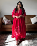 A woman modeling a bright red traditional Indian kurta set with gold embroidery and printed patterns, standing in a living room in front of a brown leather sofa.