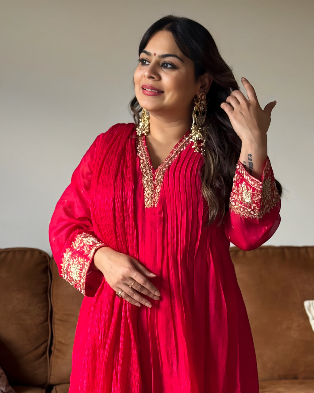 A woman wearing a bright red traditional tunic with gold embroidery on the neckline and cuffs, standing indoors in front of a neutral wall and brown sofa.