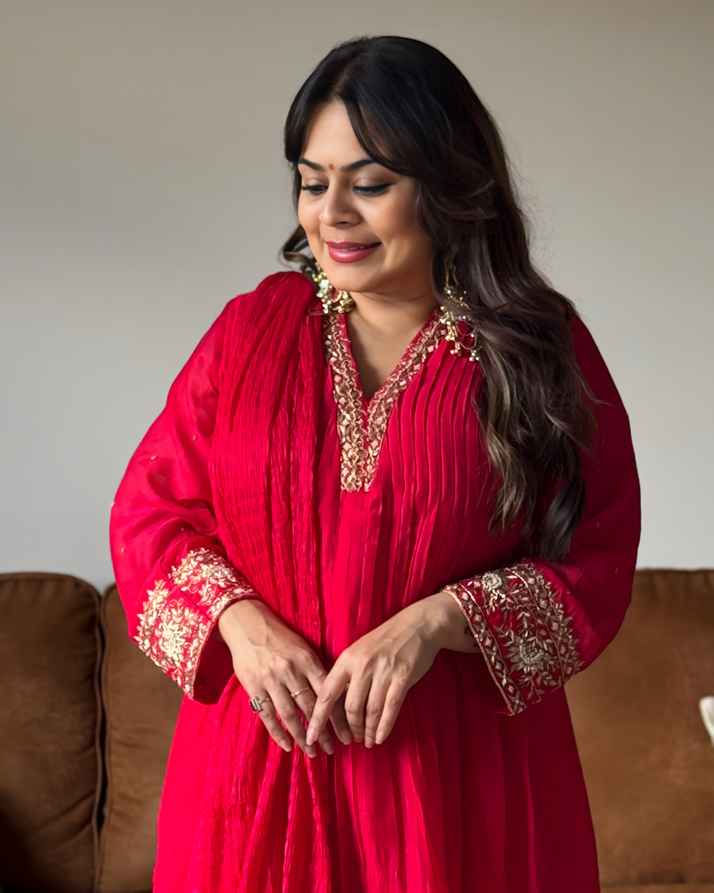 A woman wearing a bright red traditional tunic with ornate gold embroidery on the neckline, cuffs, and hem, standing indoors in front of a neutral wall and brown sofa.