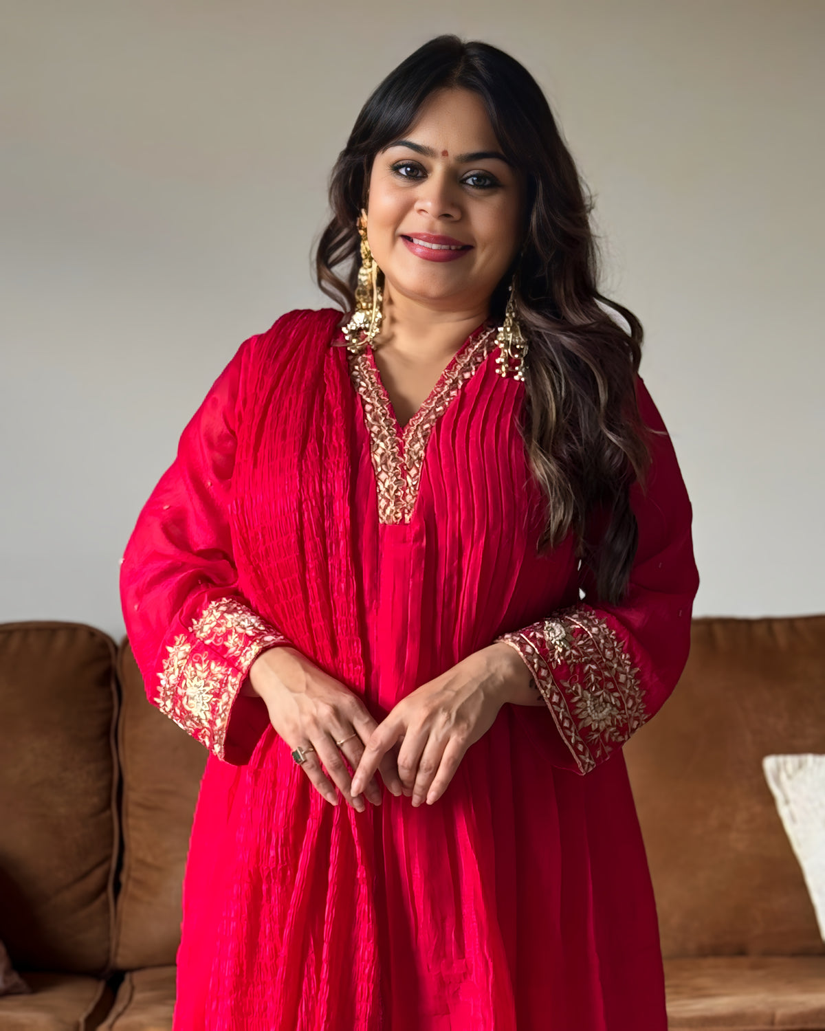 A woman wearing a bright red traditional tunic with gold embroidery on the neckline and cuffs, standing indoors in front of a brown couch against a light beige wall.