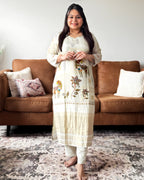 Woman in a white embroidered kurta standing in a living room with a brown sofa and patterned rug.