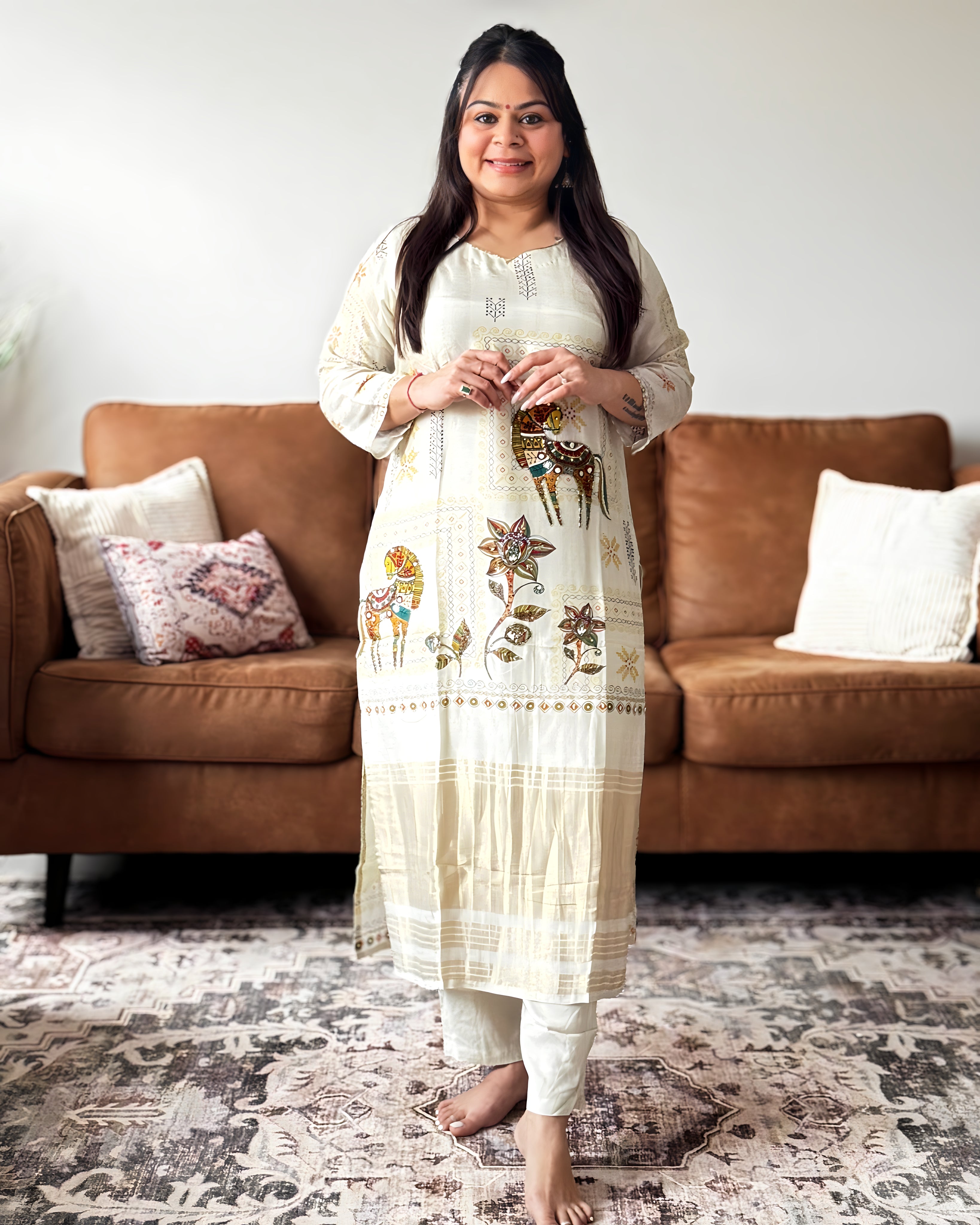 Woman in a white embroidered kurta standing in a living room with a brown sofa and patterned rug.
