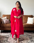 Woman in a red traditional outfit standing in a living room with a brown sofa and patterned rug.