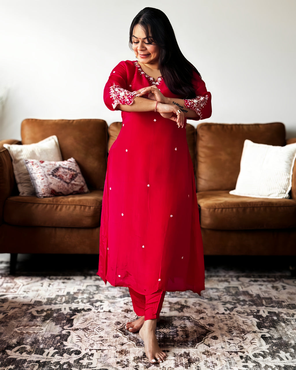 Woman in a red traditional outfit standing in a living room with a brown sofa and patterned rug.