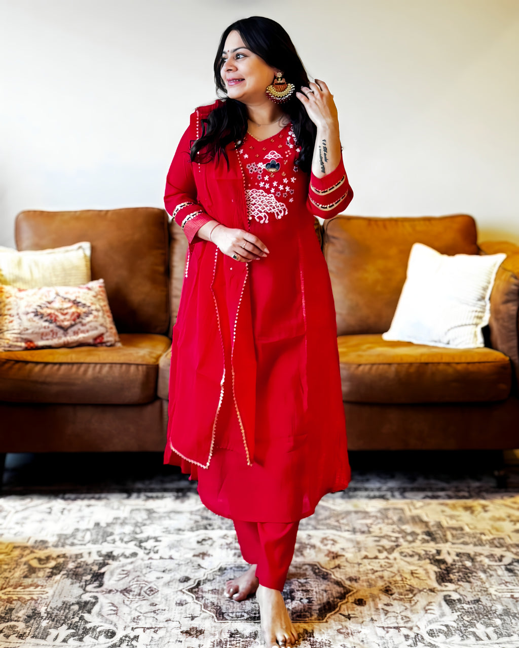 Woman in a red traditional outfit standing in a living room with a brown sofa and patterned rug.