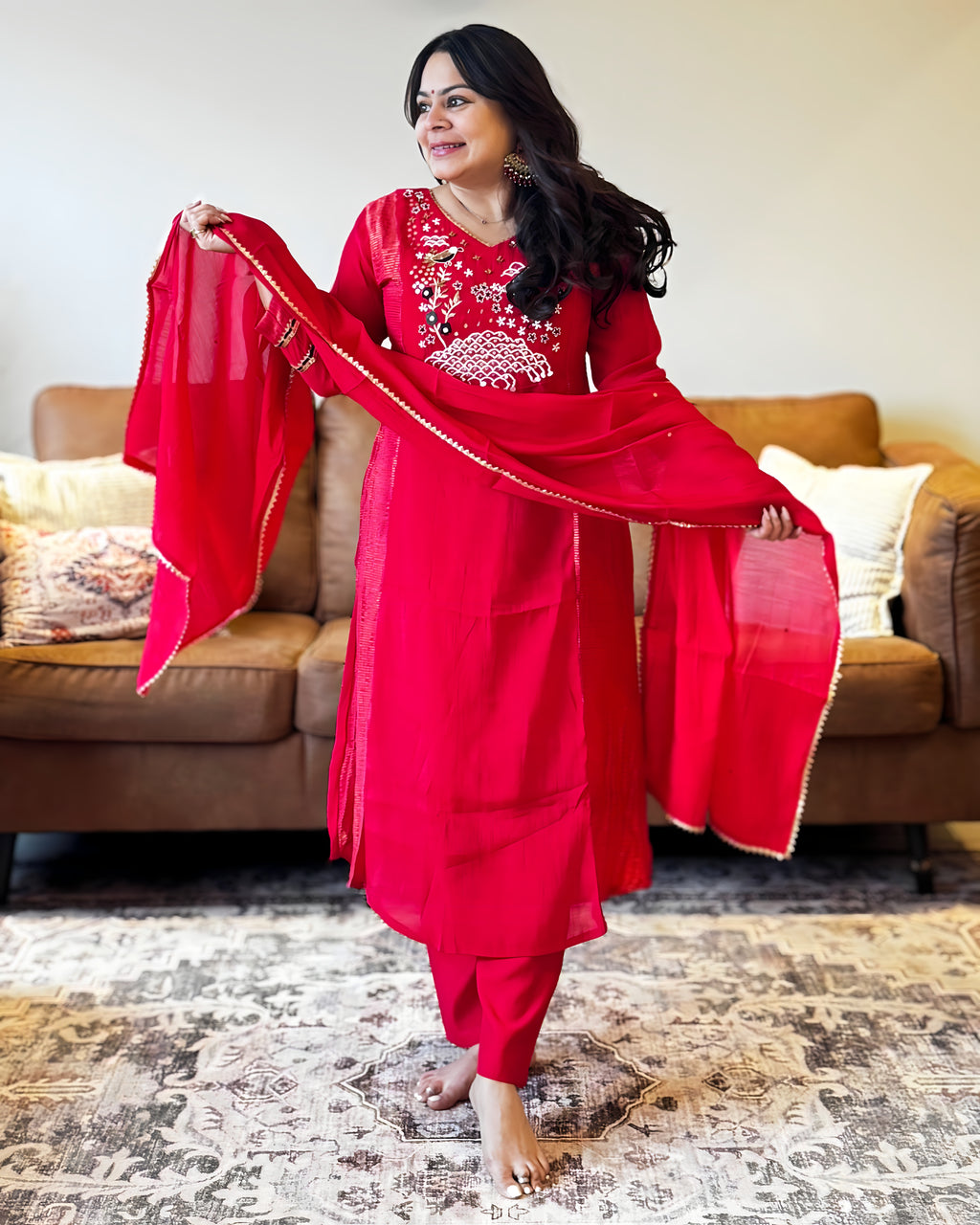 Woman in a red traditional outfit holding a dupatta in a living room.