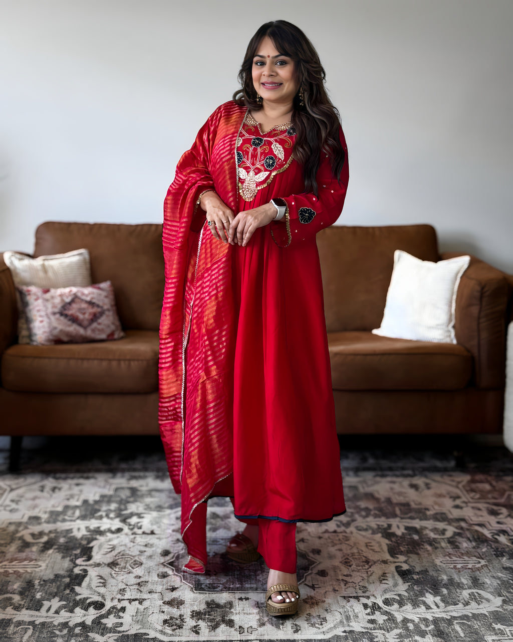A woman wearing a vibrant red traditional Indian salwar kameez suit with a matching dupatta, standing in a living room in front of a brown sofa.