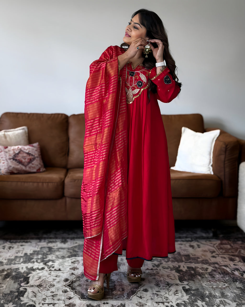 A woman models a red traditional outfit with a matching red dupatta featuring orange and pink tie-dye stripes, standing in a living room in front of a brown leather sofa.