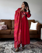 A woman models a red traditional outfit with a matching red dupatta featuring orange and pink tie-dye stripes, standing in a living room in front of a brown leather sofa.