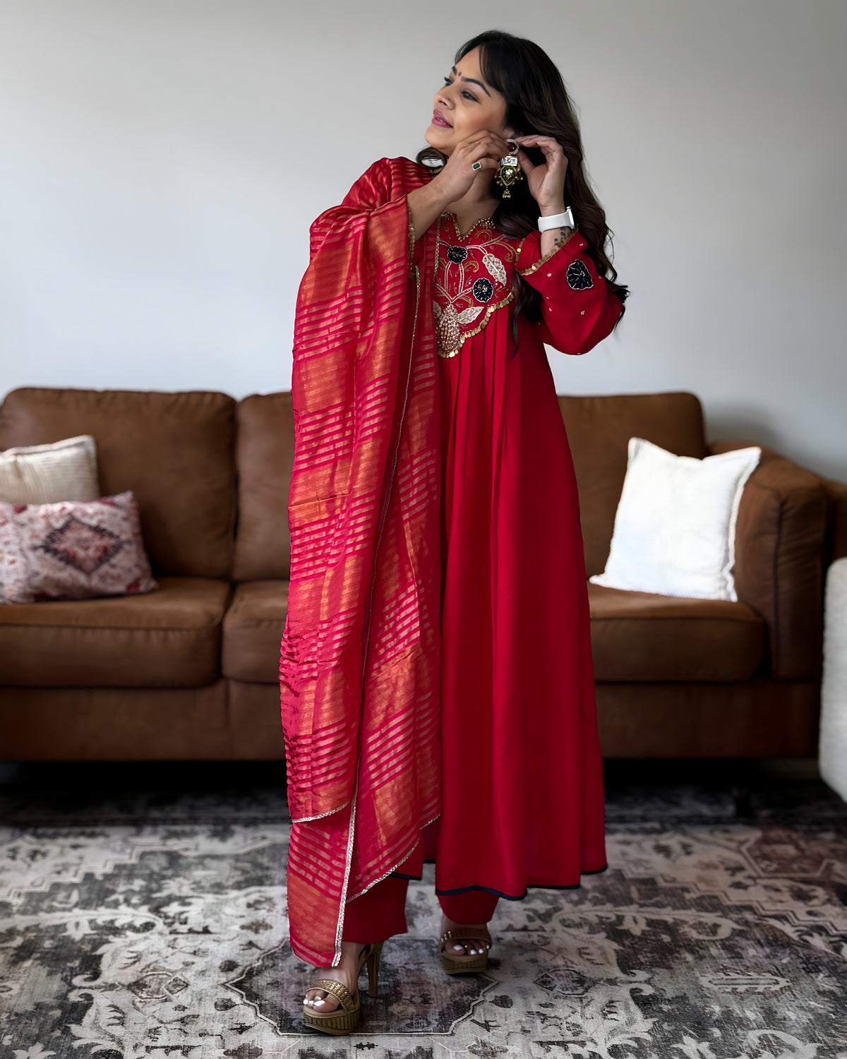 A woman models a red traditional outfit with a matching red dupatta featuring orange and pink tie-dye stripes, standing in a living room in front of a brown leather sofa.
