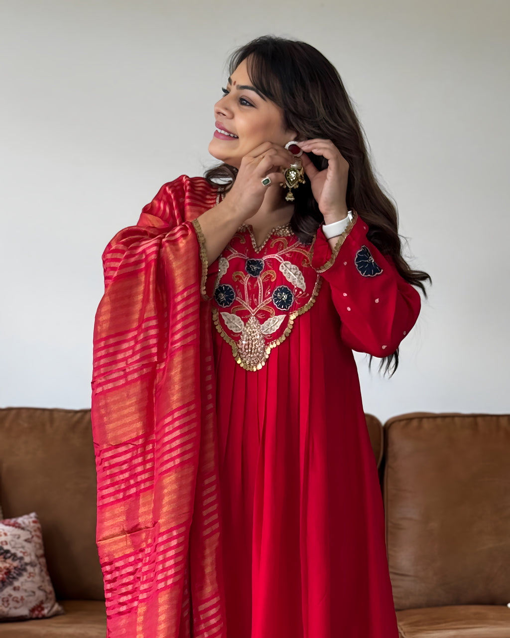 A woman wearing a vibrant red traditional outfit with intricate gold and silver embroidery on the chest, paired with a red and gold striped dupatta, standing in front of a brown leather sofa.