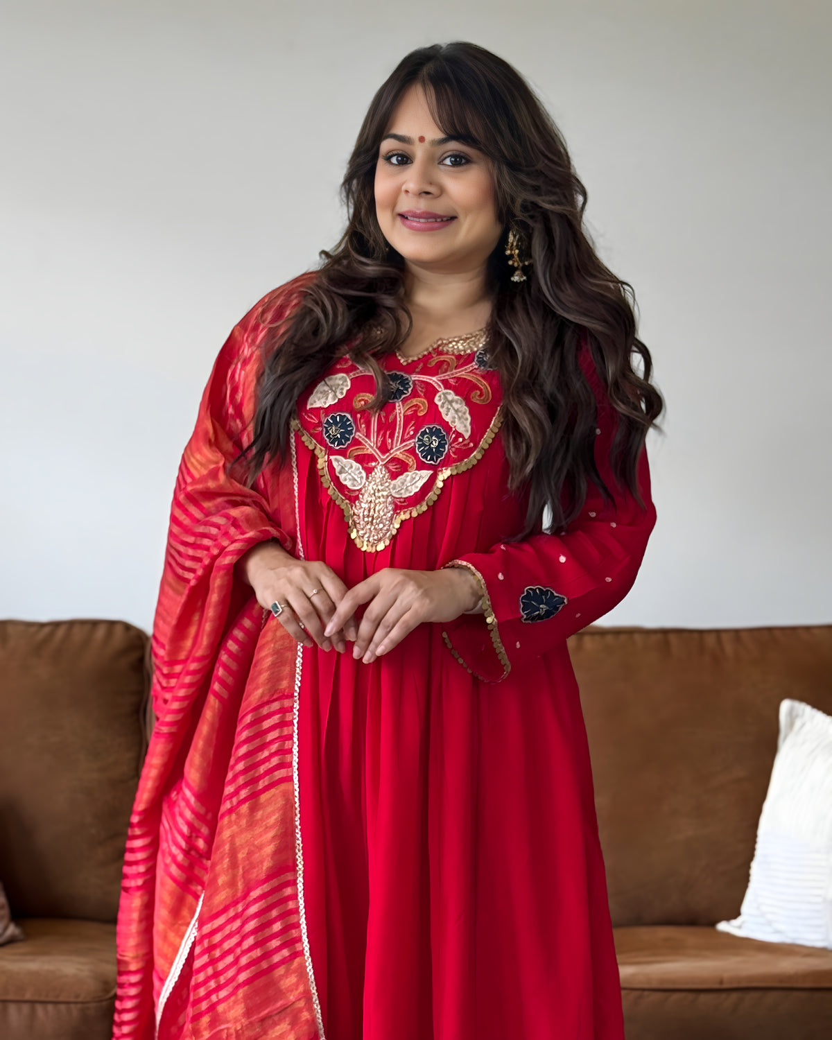 A woman wearing a vibrant red traditional Indian outfit with intricate gold and black embroidery on the chest, paired with a matching red and orange patterned dupatta, standing in front of a brown sofa.