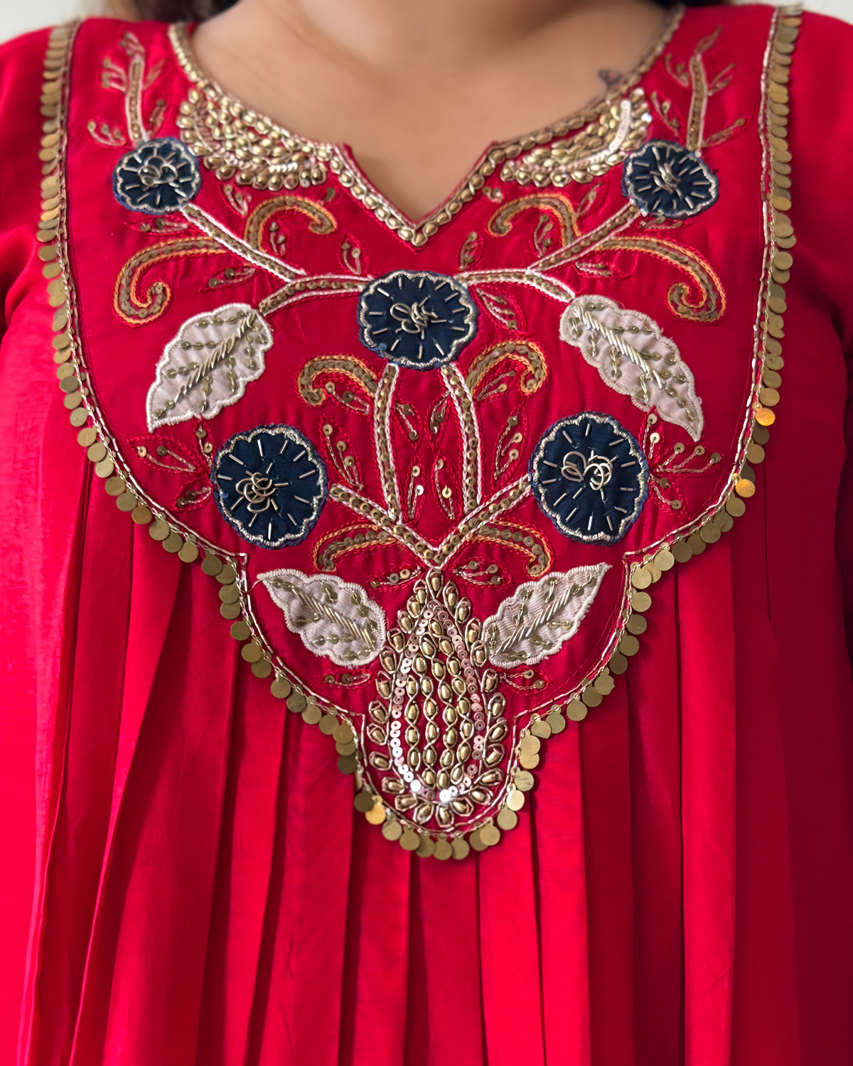 Close-up of a red embroidered garment with a V-neckline, featuring ornate floral and vine embroidery in gold, silver, and navy blue, accented with coin trim.