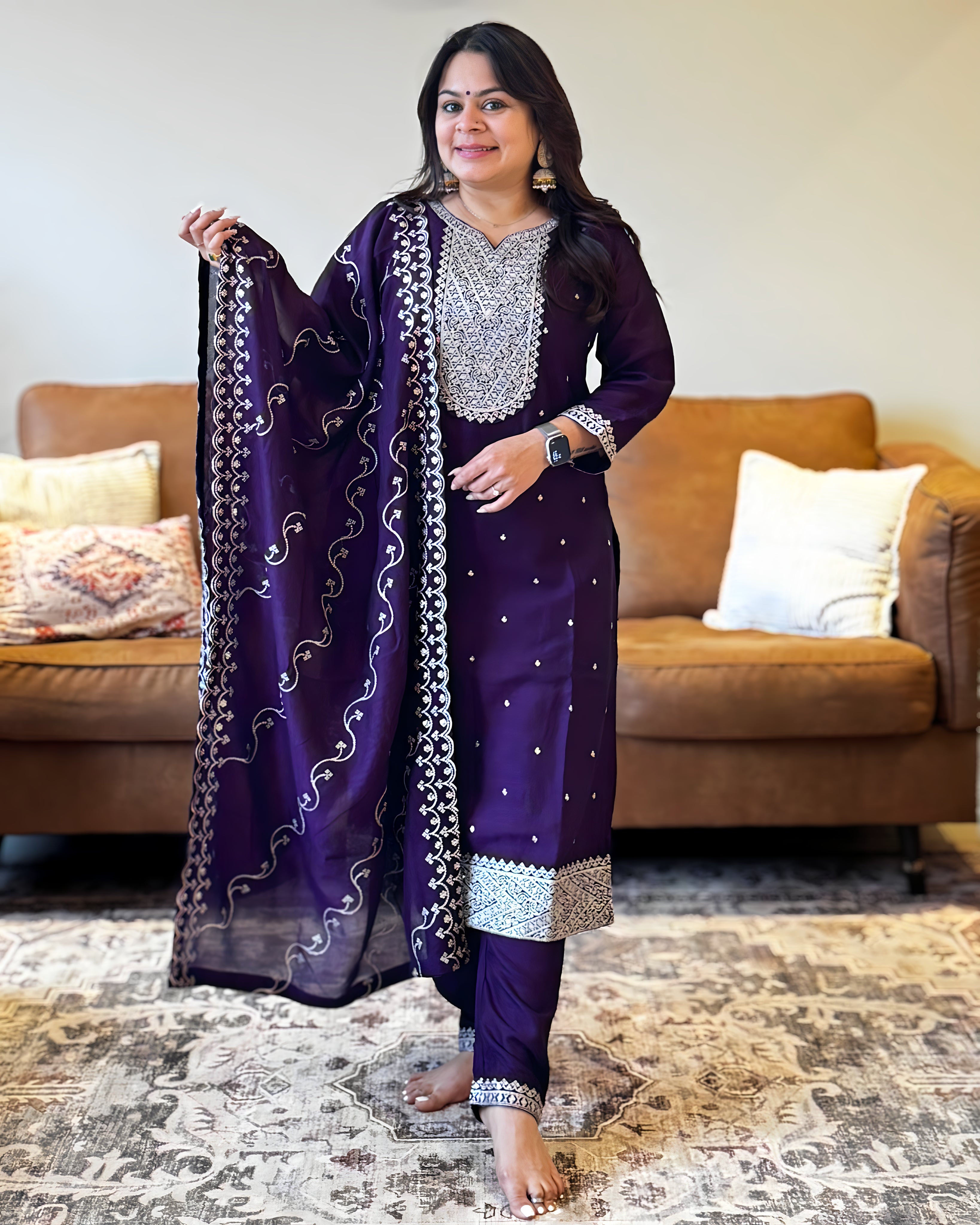 Woman in a purple traditional outfit with a matching dupatta, standing in a living room.