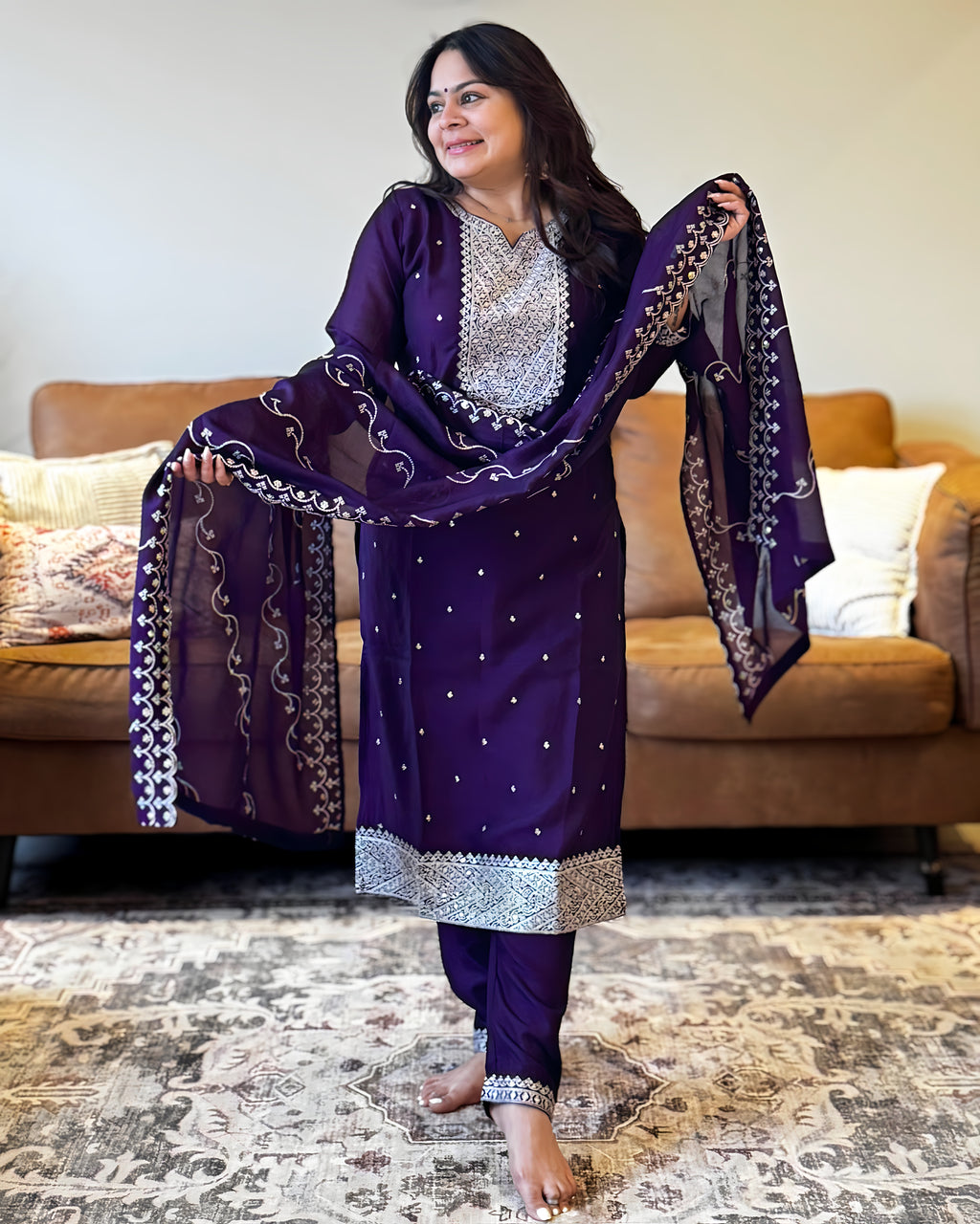 Woman in a purple traditional outfit with silver details in a living room setting.