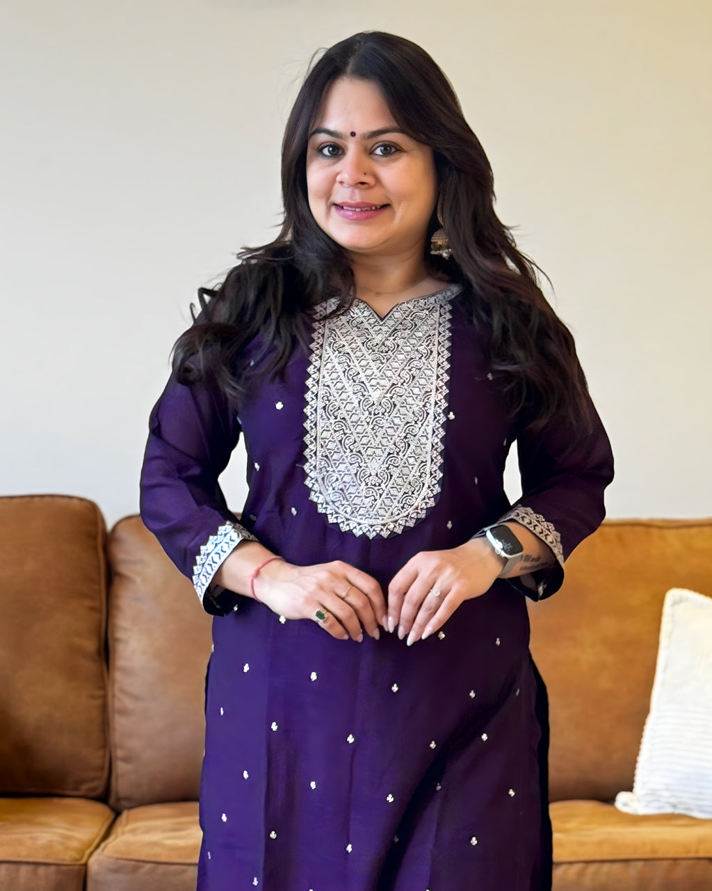 Woman wearing a purple traditional outfit with silver embroidery in a living room setting.